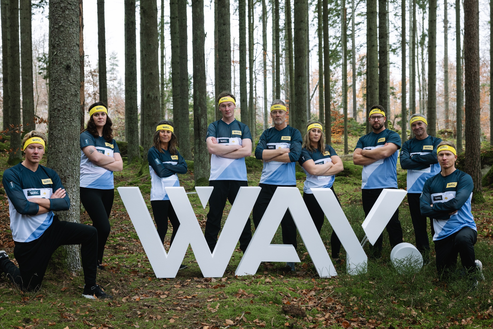 WAY-gänget står i skogen iklädda orienteringskläder runt WAY logotypen i frigolit