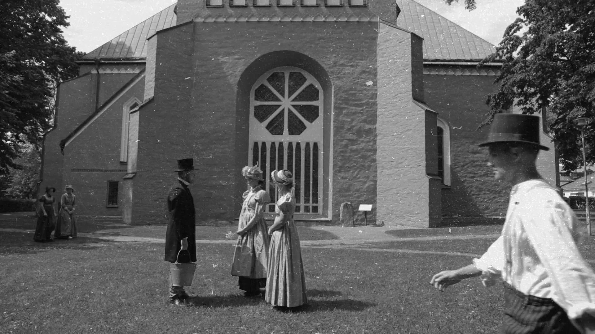 En svartvit bild på baksidan av Växjös domkyrka. Personerna i bilder står och prata och har gammeldags kläder på sig.