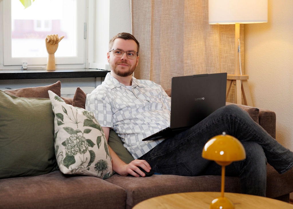 Pierre sitter och jobbar i soffan med sin laptop. Han kollar mot en kollega.