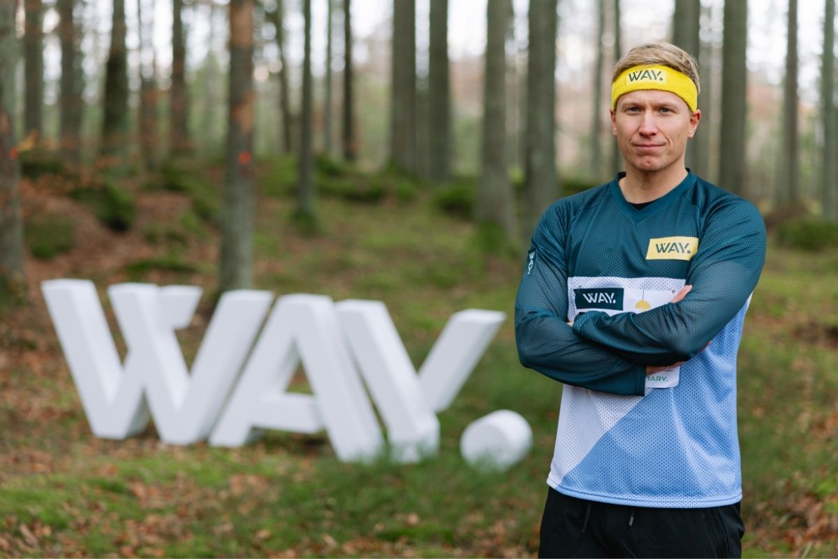 Rickard står i skogen iklädda orienteringskläder med WAY logotypen i frigolit i bakgrunden